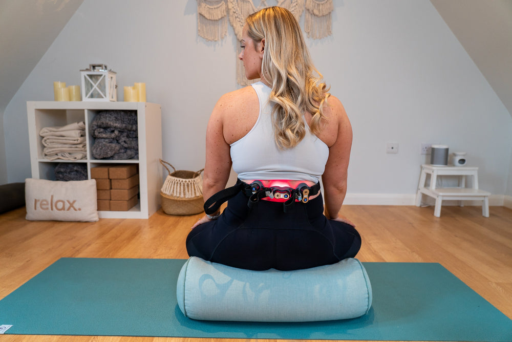 A woman using red light therapy device on her back. The device is on and red light are illuminating her lower back area.